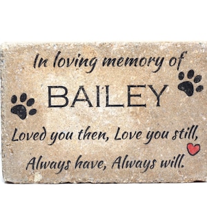 May include: A beige stone memorial with the text "In loving memory of BAILEY Loved you then, Love you still, Always have, Always will."  The stone has two black paw prints and a red heart.
