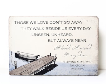 May include: A wooden sign with a quote about those we love, "Those we love don't go away. They walk beside us every day. Unseen, unheard, but always near." The sign also includes a handwritten message, "still loved, still missed & very dear." and a memorial for Jim Gavin, 1960-2019. The sign features a watercolor illustration of a dock with two boats.