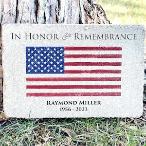 May include: A light gray stone memorial with the text "IN HONOR & REMEMBRANCE" at the top, an American flag in the center, and the text "RAYMOND MILLER 1956 - 2023" at the bottom.