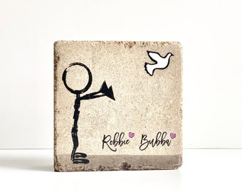 May include: A beige square tile with a black stick figure reaching out to a white dove. The tile has the text "Robbie Bubba" with two pink hearts.