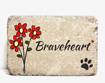 May include: A rectangular stone memorial with a hand-painted design of red flowers with yellow centers and green stems. The words "Braveheart" are written in black cursive script. A small red heart is painted next to the text. A black paw print is painted in the lower right corner.