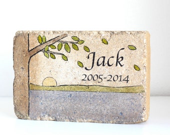 May include: A rectangular stone memorial with a hand-painted design of a tree with green leaves, a setting sun, and a grassy field. The stone is inscribed with the name "Jack" and the dates "2005-2014".