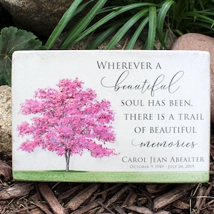May include: A white wooden sign with a pink tree and the text "WHEREVER A beautiful SOUL HAS BEEN, THERE IS A TRAIL OF BEAUTIFUL memories. CAROL JEAN ABFALTER OCTOBER 9, 1949 - JULY 24, 2019"