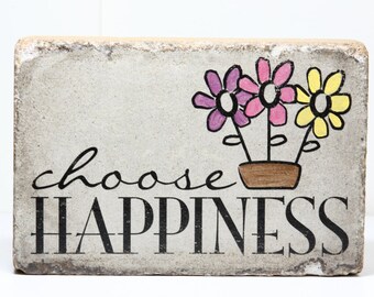 May include: A decorative concrete block with the words "choose happiness" in black lettering. The block is decorated with a hand-drawn image of three flowers in a brown pot.