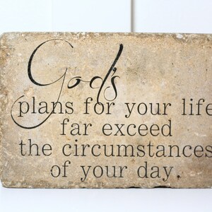 May include: A beige stone with the text "God's plans for your life far exceed the circumstances of your day." printed in black.