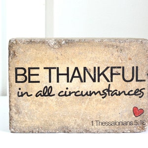 May include: A beige stone block with the text "BE THANKFUL in all circumstances" and a small red heart. The bottom of the block has the text "1 Thessalonians 5:18".