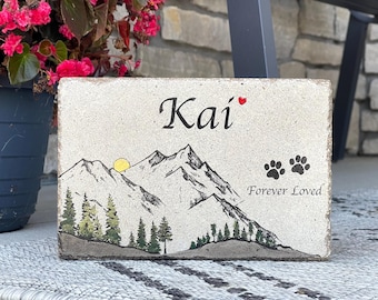 May include: A light beige stone memorial with a black mountain range silhouette, a yellow sun, and green trees. The name "Kai" is written in black with a red heart. The words "Forever Loved" are written in black with two black paw prints above them.