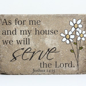 May include: A rectangular stone plaque with the inscription "As for me and my house we will serve the Lord." The text is in black, with a floral design of white daisies with yellow centers. The plaque is a light brown color.