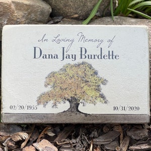 May include: A rectangular memorial stone with the inscription "In Loving Memory of Dana Jay Burdette." The stone features a tree illustration and the dates 02/20/1955 and 10/31/2020. The stone is light-colored with a textured surface.