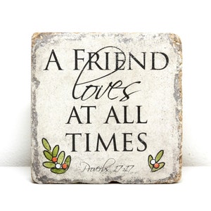 May include: A white stone coaster with the text "A friend loves at all times" and the scripture reference "Proverbs 17:17". The coaster has a rustic, distressed look and features green leaves and red berries.