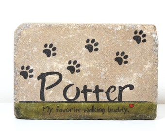 May include: A light brown stone with black paw prints and the name "Potter" written in black cursive. The stone has a green band at the bottom with the text "My favorite walking buddy." in black cursive and a red heart.