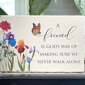 May include: A white wooden sign with a floral design and a butterfly. The sign reads "A friend is God's way of making sure we never walk alone."