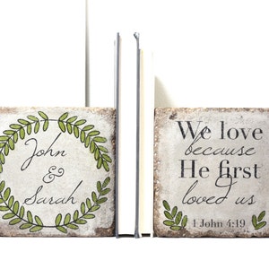 May include: A set of two bookends made of stone with a white background. One bookend features a green leafy wreath with the names "John & Sarah" inside. The other bookend features the text "We love because He first loved us 1 John 4:19" with a green leafy design at the bottom.