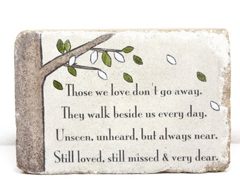 May include: A white stone with a painted tree branch and leaves. The stone has a quote about remembering loved ones: "Those we love don't go away. They walk beside us every day. Unseen, unheard, but always near. Still loved, still missed & very dear."