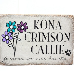 May include: A white stone block with the names "Kona Crimson Callie" and the text "forever in our hearts" written in black. The block is decorated with a hand-painted bouquet of purple, teal, and white flowers.