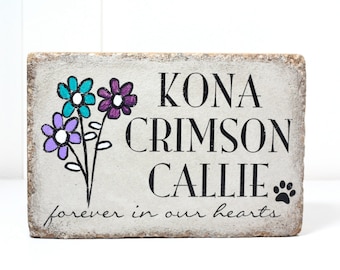 May include: A white stone block with the names "Kona Crimson Callie" and the text "forever in our hearts" written in black. The block is decorated with a hand-painted bouquet of purple, teal, and white flowers.