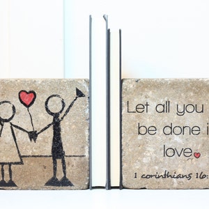 May include: A pair of bookends made of concrete with a hand-painted design. One bookend features a stick figure couple holding a red heart balloon. The other bookend has the text "Let all you do be done in love" with a small red heart and the scripture reference "1 Corinthians 16:14".