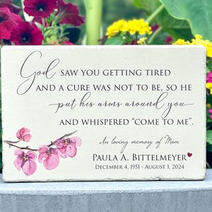 May include: A rectangular memorial plaque with a cream-colored background. The plaque features elegant script with the words "God saw you getting tired" and a heartfelt message. Pink floral accents and the name Paula A. Bittelmeyer are also included.