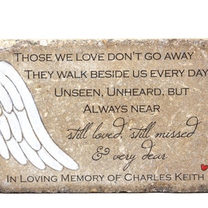 May include: A beige stone memorial with a white painted angel wing design. The stone has the text "Those we love don't go away. They walk beside us every day. Unseen, unheard, but always near. Still loved, still missed & very dear. In loving memory of Charles Keith."