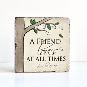 May include: A square wooden sign with a beige background and a painted tree branch with green leaves and white blossoms. The sign reads "A friend loves at all times" with the scripture reference "Proverbs 17:17" below.