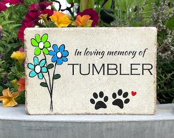May include: A light beige stone memorial with the text "In loving memory of TUMBLER" and two black paw prints. The stone is decorated with three painted flowers in shades of blue, green, and white.