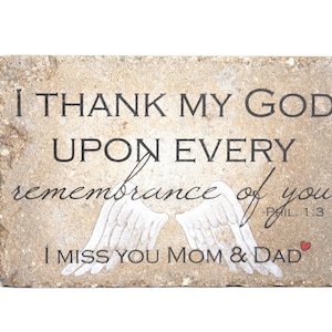 May include: A stone plaque with a white background and black text that reads "I thank my God upon every remembrance of you - Phil. 1:3".  The plaque also features a pair of white angel wings and the text "I miss you Mom & Dad" with a small red heart.