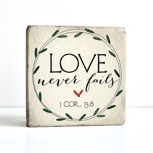 May include: A square, off-white wooden block with the words "LOVE never fails" in black script, encircled by a green leaf wreath. A red heart and the text "1 COR. 13:8" are also present. The block has a slightly distressed appearance.