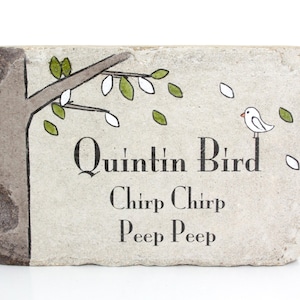 May include: A white stone with a hand-painted design of a tree branch with green leaves, a white bird, and the text "Quintin Bird Chirp Chirp Peep Peep".