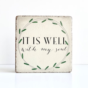 May include: A white wooden sign with a green wreath around the words "It is well with my soul".