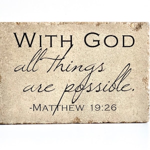 May include: A rectangular, beige stone block with the words "WITH GOD all things are possible." in black script. The text "-MATTHEW 19:26" is below. The stone has a rough, textured surface.