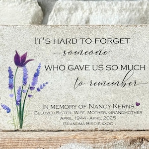 May include: A rectangular stone memorial with the text "IT'S HARD TO FORGET someone WHO GAVE US SO MUCH to remember." A purple flower and lavender sprigs are on the left. The memorial is "IN MEMORY OF NANCY KERNS".