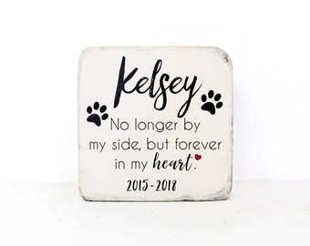 May include: White wooden sign with black text that reads "Kelsey No longer by my side, but forever in my heart. 2015-2018". There are two black paw prints above the name and a small red heart after the word "heart".
