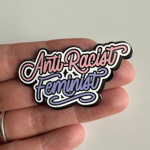 May include: A pin with the words "Anti-Racist Feminist" in a stylized script. The text is in pink and purple with a black outline and white accents. The pin is held in a hand.
