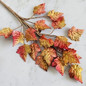 May include: A decorative branch featuring multiple artificial maple leaves in a gradient of metallic gold, orange, and deep red. The leaves are attached to a thin, brown, textured stem that branches out to support the foliage in a natural arrangement.