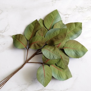 May include: A bunch of artificial green leaves with red veins, arranged on a white marble background.
