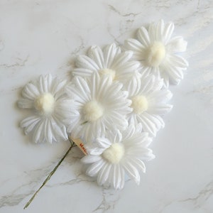 May include: A cluster of white daisy flowers with yellow centers and green stems. The flowers are arranged on a white marble surface. A small tag is attached to the stem. The flowers are a popular choice for floral arrangements and crafts.