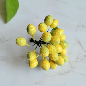 May include: A bunch of small, yellow and green berry-like floral picks. The berries are clustered together on a green stem.