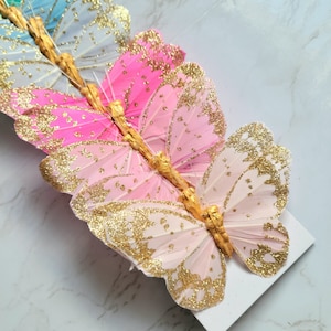 May include: A set of six pink, blue, and green butterfly decorations with gold glitter accents. The butterflies are made of feathers and have a wire attached to the back for hanging.