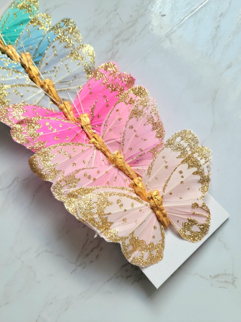 May include: A set of six pink, blue, and green butterfly decorations with gold glitter accents. The butterflies are made of feathers and have a wire attached to the back for hanging.