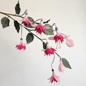 May include: A decorative branch featuring multiple fuchsia flowers with light pink outer petals and deep magenta inner petals. The flowers hang from a thin brown stem adorned with textured dark green leaves. Each bloom has a small blue-grey base.