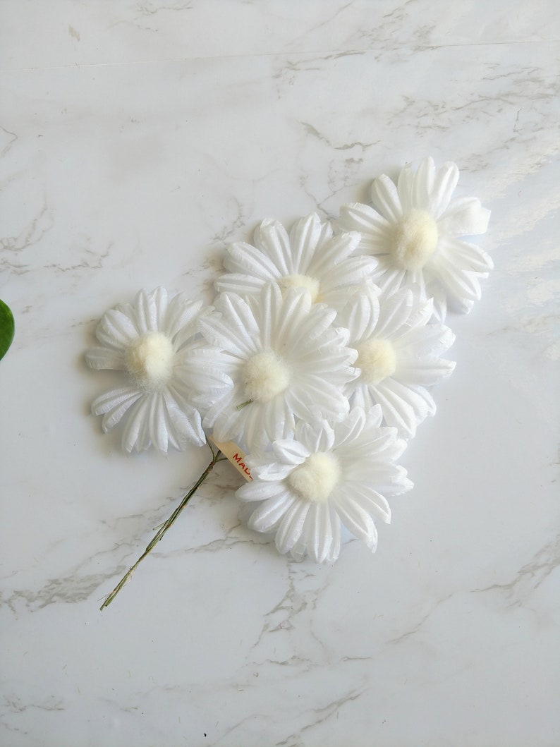 May include: A cluster of white daisy flowers with yellow centers and green stems. The flowers are arranged on a white marble surface. A small tag is attached to the stem. The flowers are a popular choice for floral arrangements and crafts.