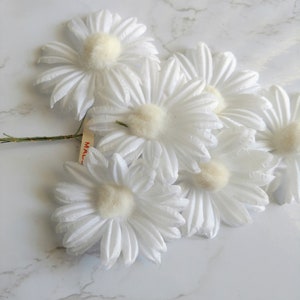 May include: A cluster of white daisy flowers with soft, fluffy centers and delicate petals. The daisies are arranged on a green stem, with a small tag attached. The flowers are set against a white marble background.