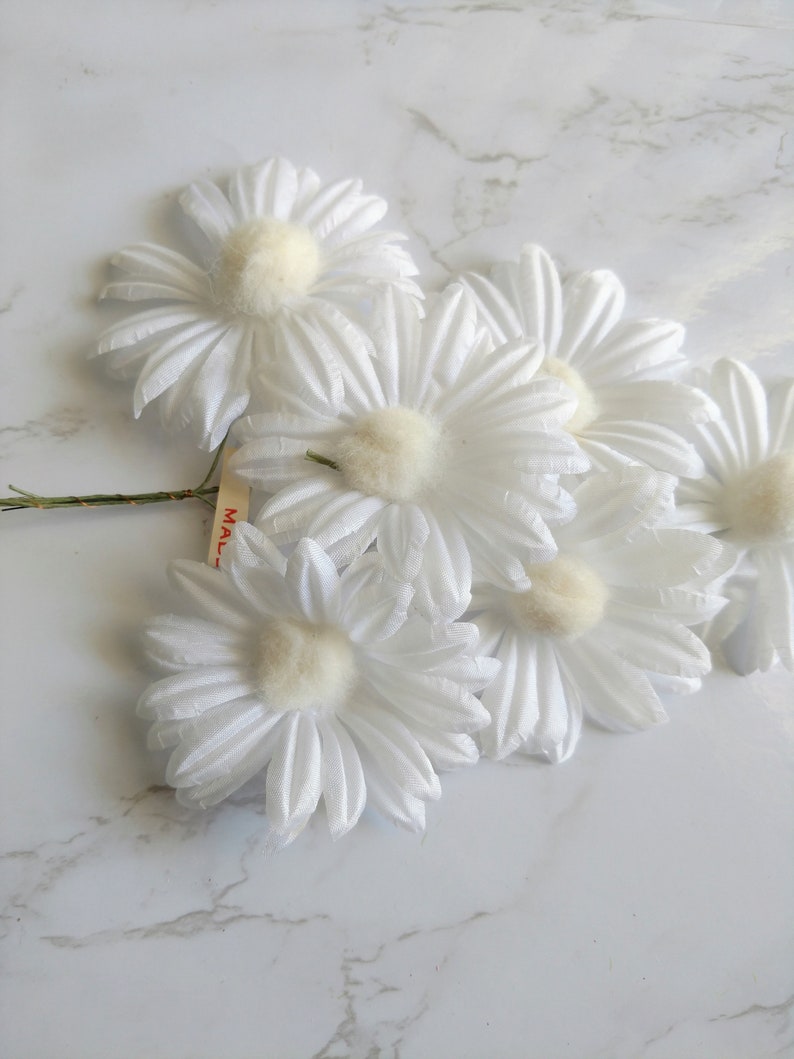 May include: A cluster of white daisy flowers with soft, fluffy centers and delicate petals. The daisies are arranged on a green stem, with a small tag attached. The flowers are set against a white marble background.