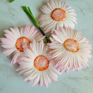 Puede incluir: Un ramo de cuatro margaritas artificiales de color rosa con centros amarillos y motas rojas. Las margaritas est&aacute;n dispuestas en un racimo con sus tallos entrelazados.