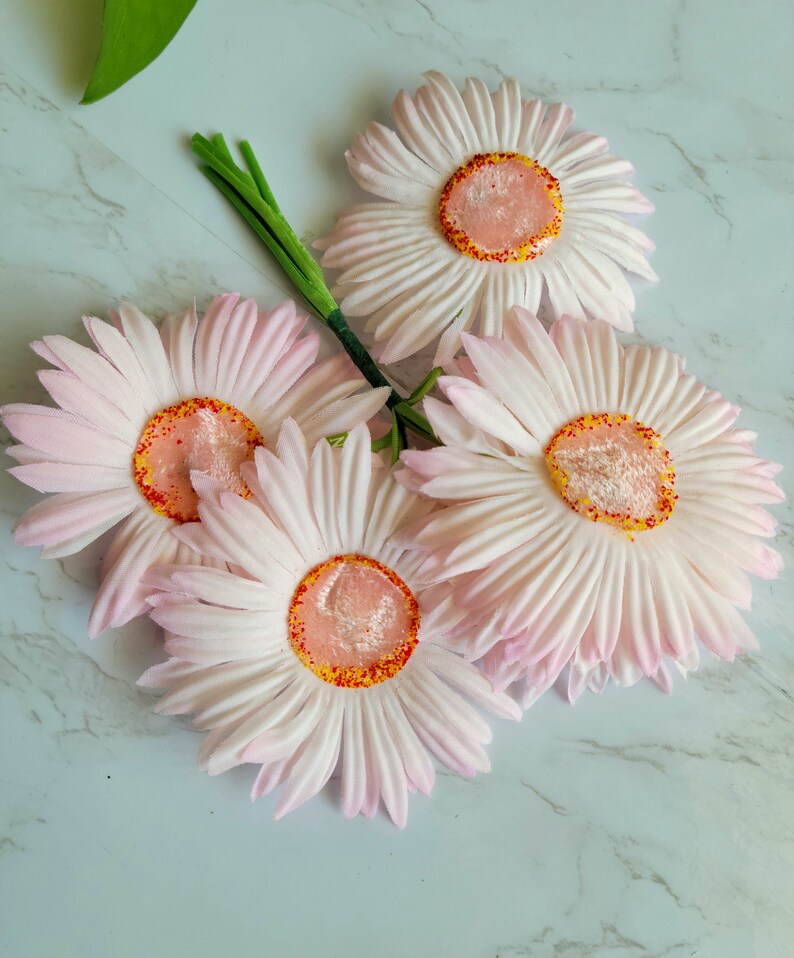 Puede incluir: Un ramo de cuatro margaritas artificiales de color rosa con centros amarillos y motas rojas. Las margaritas est&aacute;n dispuestas en un racimo con sus tallos entrelazados.
