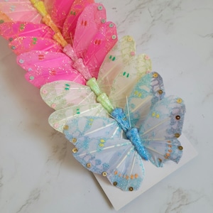 May include: A set of colorful butterfly hair clips with glitter and sequins. The butterflies are arranged in a rainbow pattern, starting with pink and ending with blue.