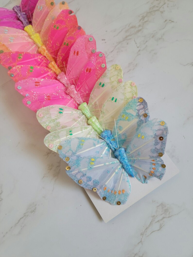 May include: A set of colorful butterfly hair clips with glitter and sequins. The butterflies are arranged in a rainbow pattern, starting with pink and ending with blue.