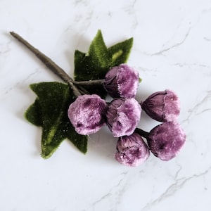 May include: A decorative floral sprig featuring five small, rounded, purple velvet-covered buds clustered together. The buds are attached to a thin, dark brown stem and are accented by two pointed, dark green velvet leaves.