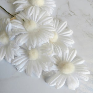 May include: A cluster of white daisy flowers with soft, textured petals and a fuzzy, cream-colored center. The flowers are arranged on a light-colored surface, creating a delicate and natural aesthetic. The stems are visible, suggesting they are artificial.