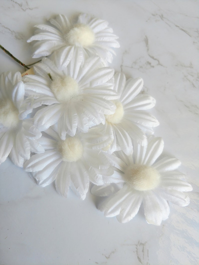 May include: A cluster of white daisy flowers with soft, textured petals and a fuzzy, cream-colored center. The flowers are arranged on a light-colored surface, creating a delicate and natural aesthetic. The stems are visible, suggesting they are artificial.